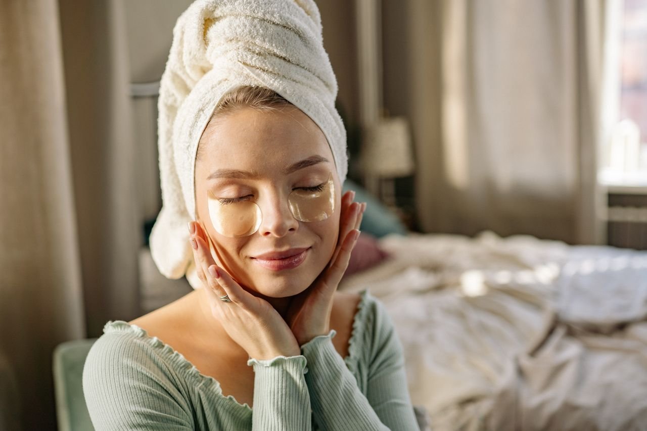 Woman enjoying a relaxing skincare routine with eye patches and towel at home.